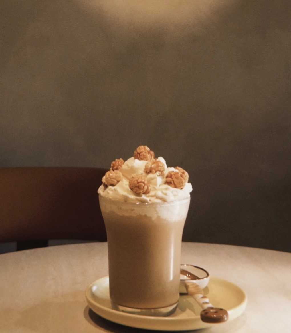 OmO Cafe 歐莫爆米花奶油拿鐵，溫暖微光下的玻璃杯特調咖啡，頂部擠滿滑順奶油與飽滿爆米花顆粒